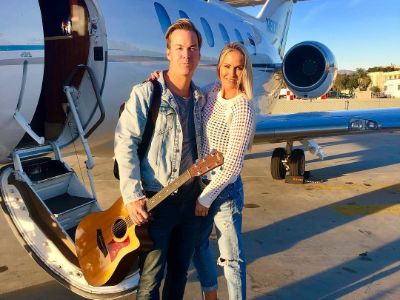 Jaclyn Dahm and Billy Dolan are posing outside the airplane as Billy is holding the guitar.
